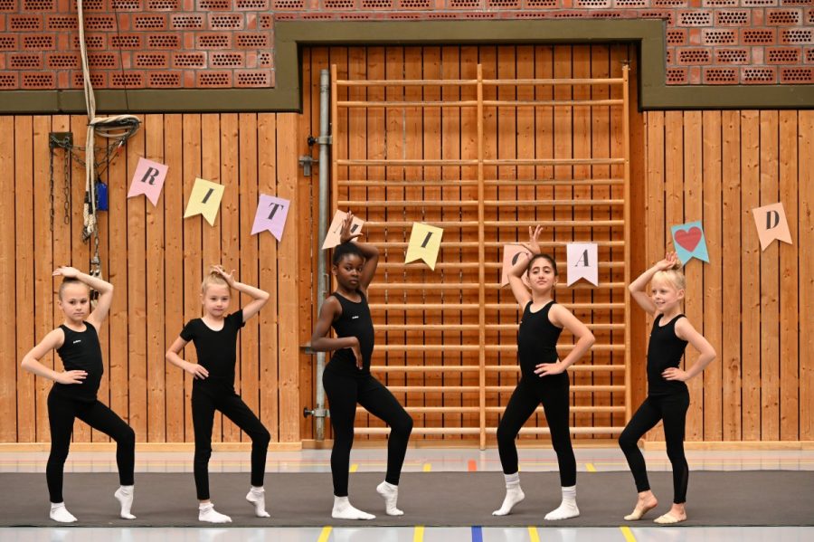 Fünf Gymnastinnen stehen nebeneinander in einer Sporthalle und posieren.