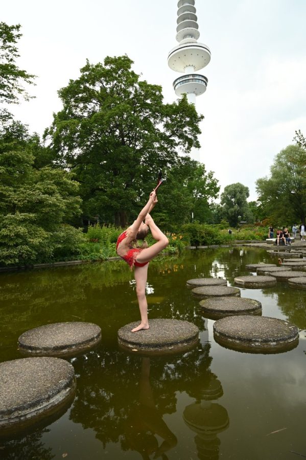 Eine Gymnastin zeigt eine Standwaage mit Bogen auf Trittsteinen in einem Teich vor einem Fernsehturm.