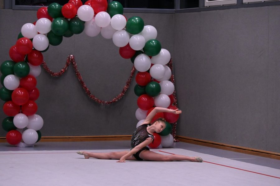 Eine Gymnastin zeigt einen Spagat auf einer Matte in einer Halle vor einem Ballonbogen.