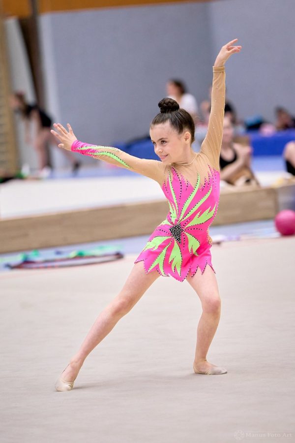 Eine Gymnastin steht in der Sporthalle auf der Wettkampffläche und breitet die Arme aus.