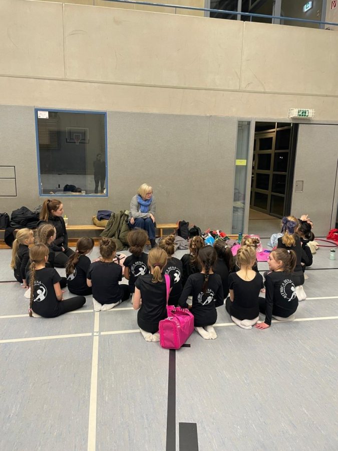 Eine Gruppe Kinder sitzt in einer Turnhalle auf dem Hallenboden vor Erwachsenen.