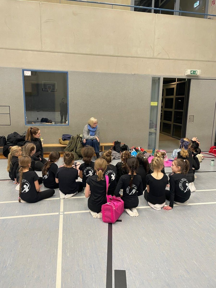 Eine Gruppe Kinder sitzt in einer Turnhalle auf dem Hallenboden vor Erwachsenen.