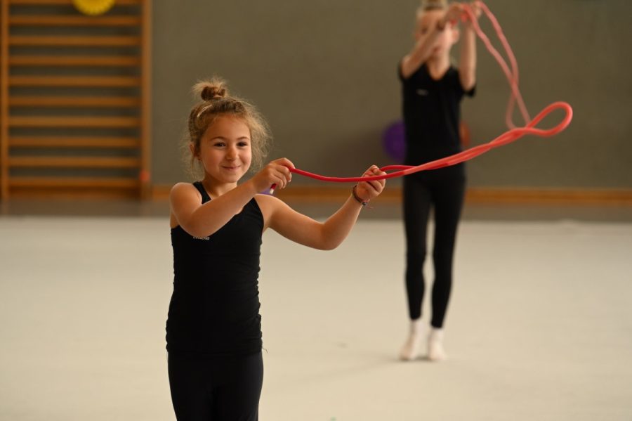 Ein Mädchen lächelt beim Training mit einem Seil in der Turnhalle, im Hintergrund eine weitere Gymnastin.