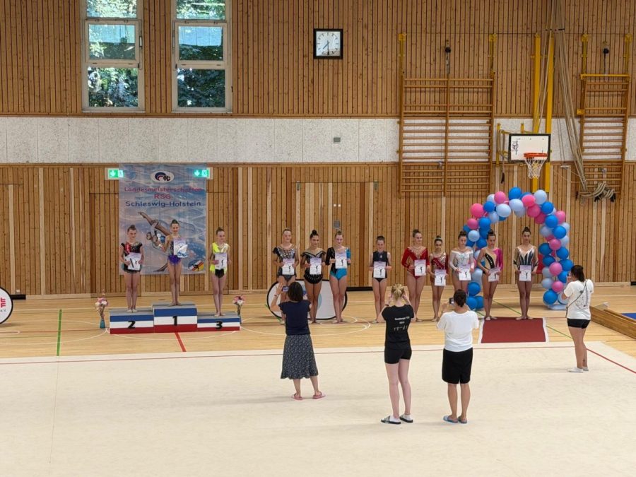 Several gymnasts stand on and beside podiums during an award ceremony in a sports hall.